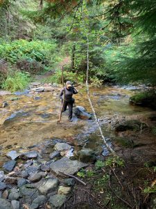a person crosses a creek using a tall walking stick