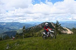 a motocross biker dressed in red rides up a grassy hill, overlooking mountains