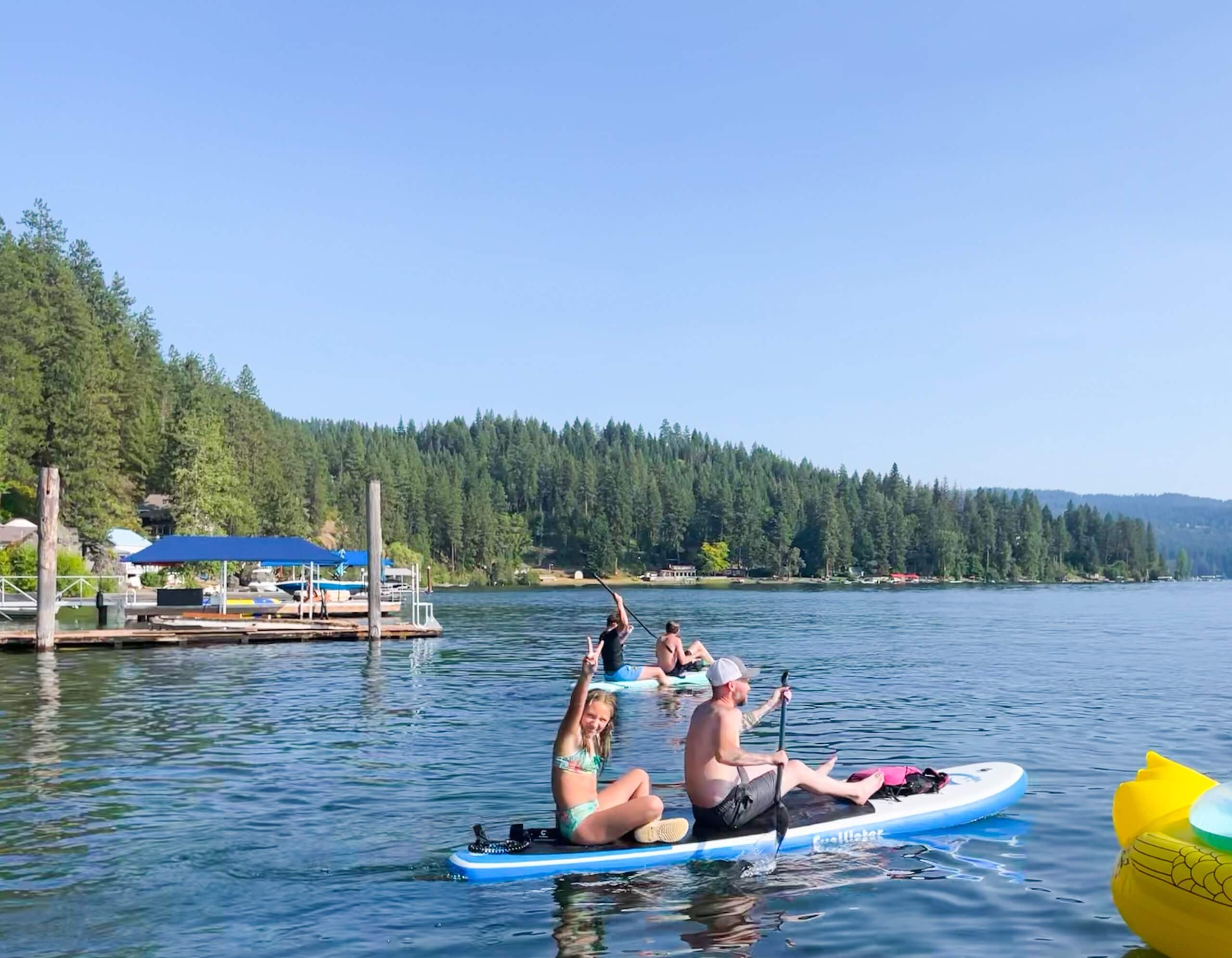 Groups of people paddleboarding near the marina on Lake Coeur d'Alene and a thick forest of trees along the shoreline in the background.