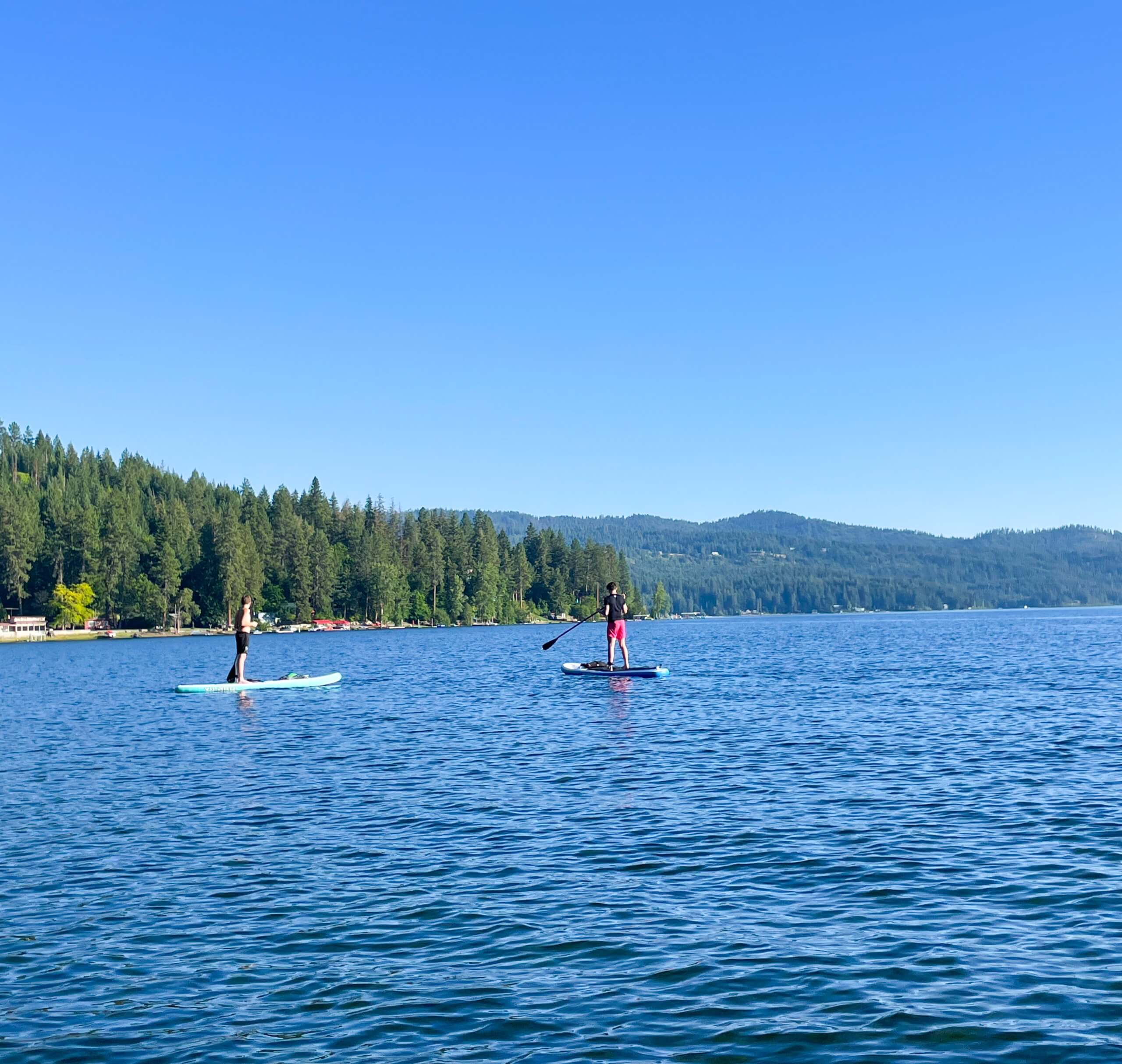 Two people paddleboarding on Spirit Lake in front of a boat dock, and a shore lined with trees and houses in the background.