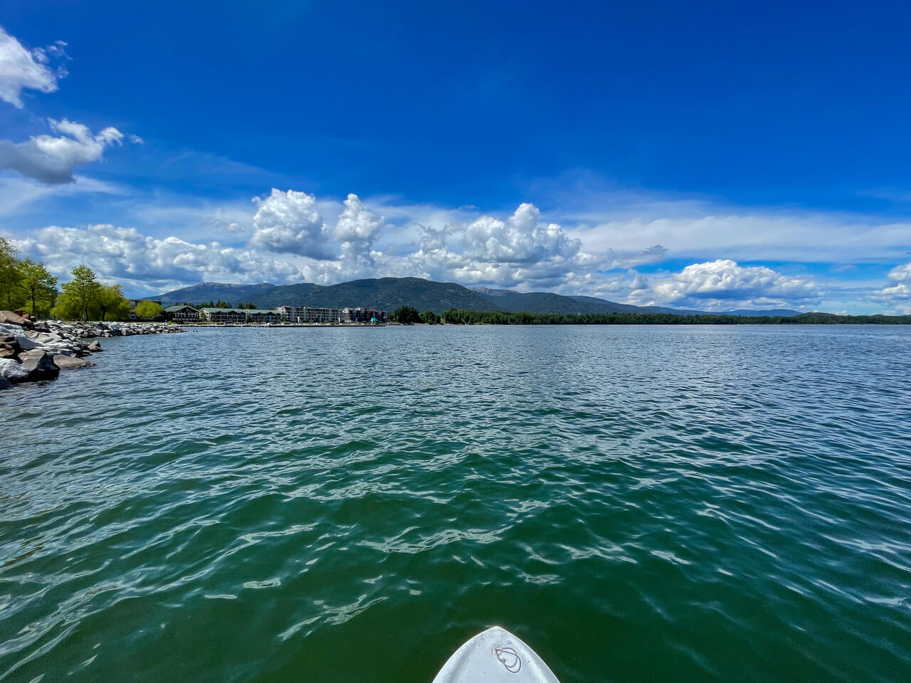 The tip of a paddleboard in the foreground, and before it, the expansive waters of Lake Pend Oreille, lined by a rocky shore.