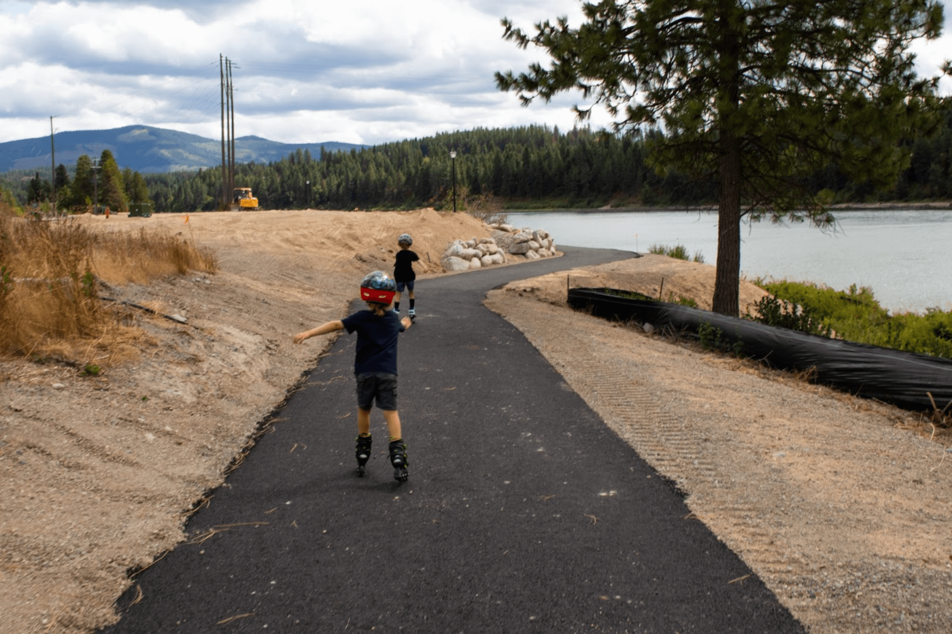 Two children rollerblading down the paved Pend Oreille Passage Trail past a river, and a forest of trees in the distance.