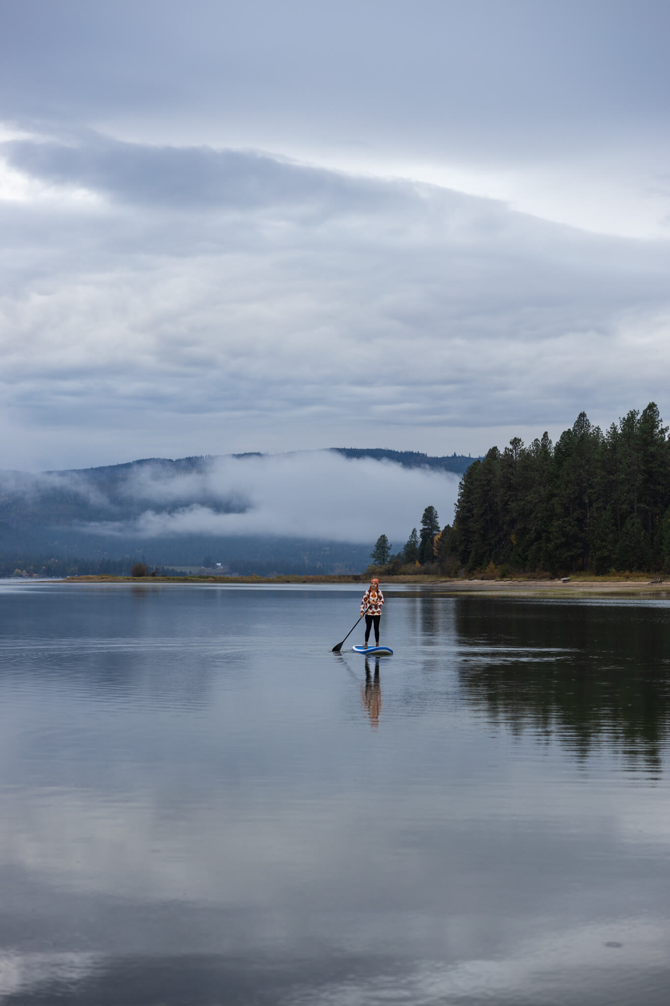 A person paddleboarding on the tree-lined Pend Oreille River at Riley Creek Recreation Area on a foggy day.