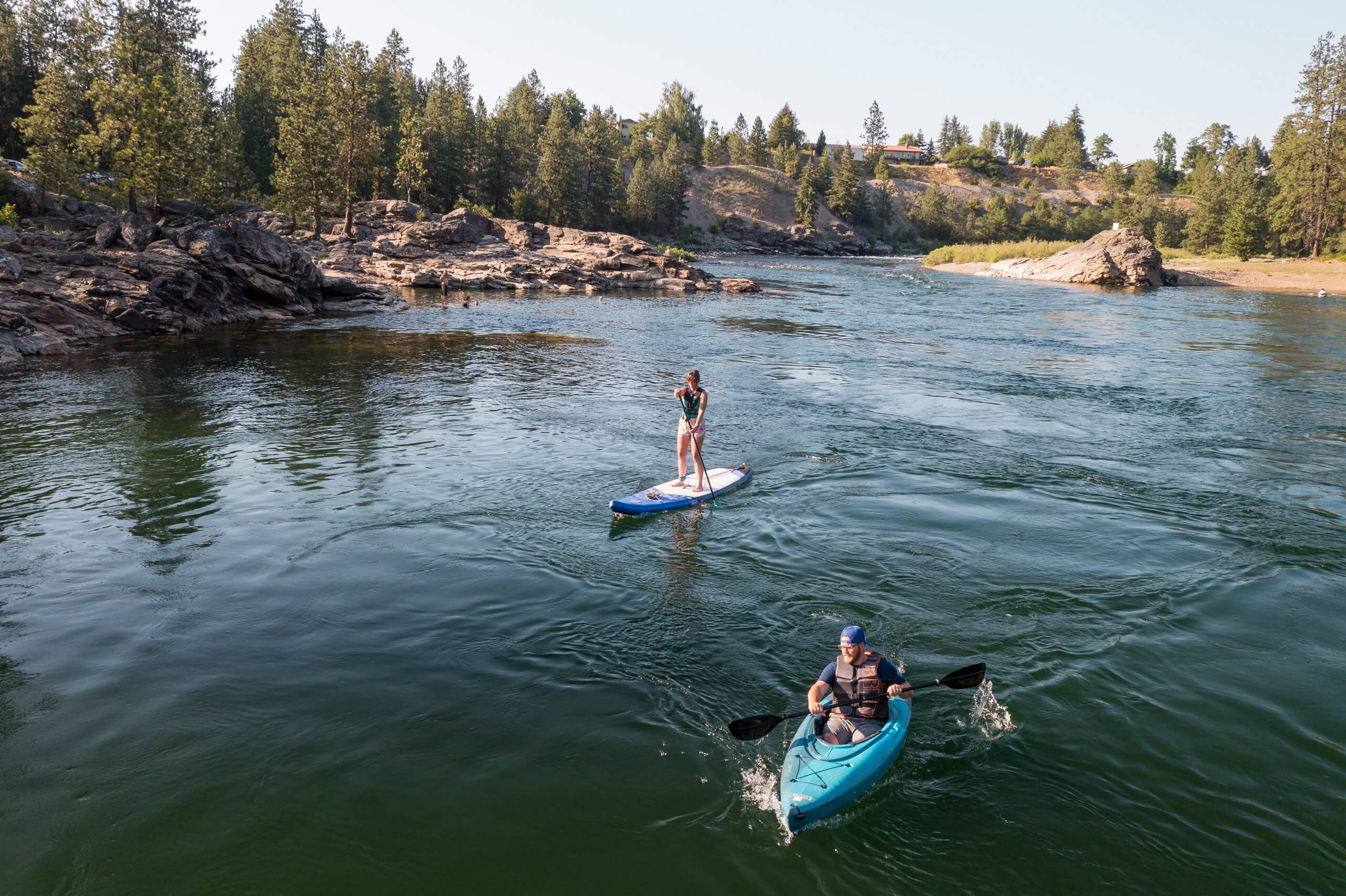 A person paddleboarding and a person kayaking on the Spokane River, surrounded by rocky shores and tall trees.