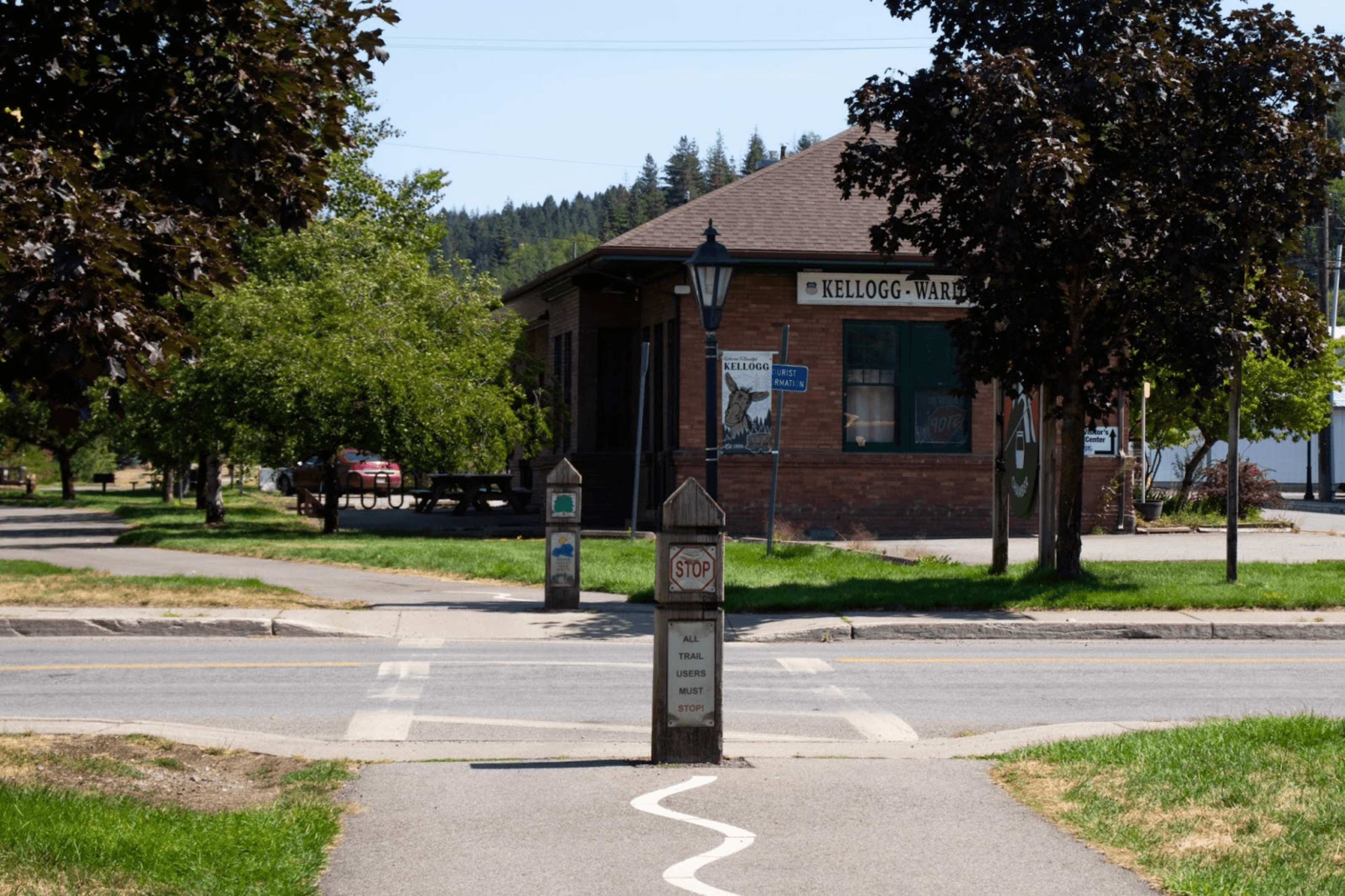 The paved Trail of the Coeur d'Alenes leading through a field of grass, across a street and past a small brick building and groups of trees.