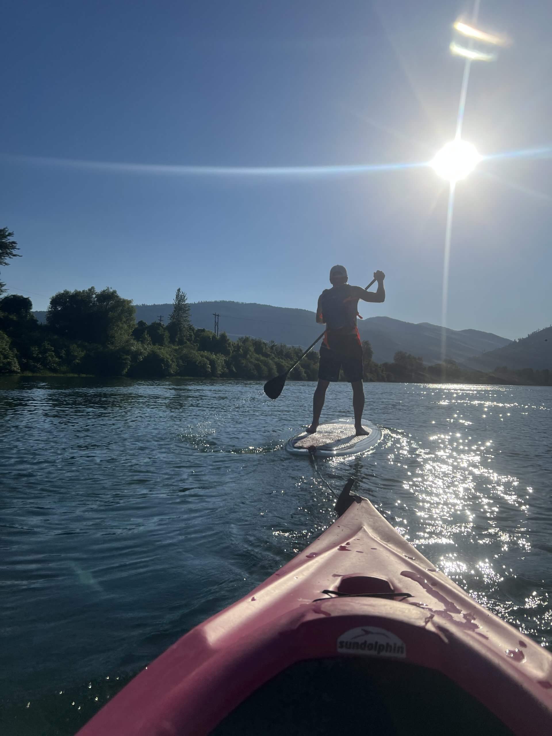 The tip of a kayak in the foreground and a person paddleboarding up ahead on the tree-lined Kootenai River.