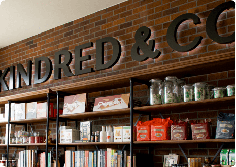 A backlit sign against a brick wall that reads Kindred & Co., and below it, multiple shelves with books and various products at Kindred & Co.