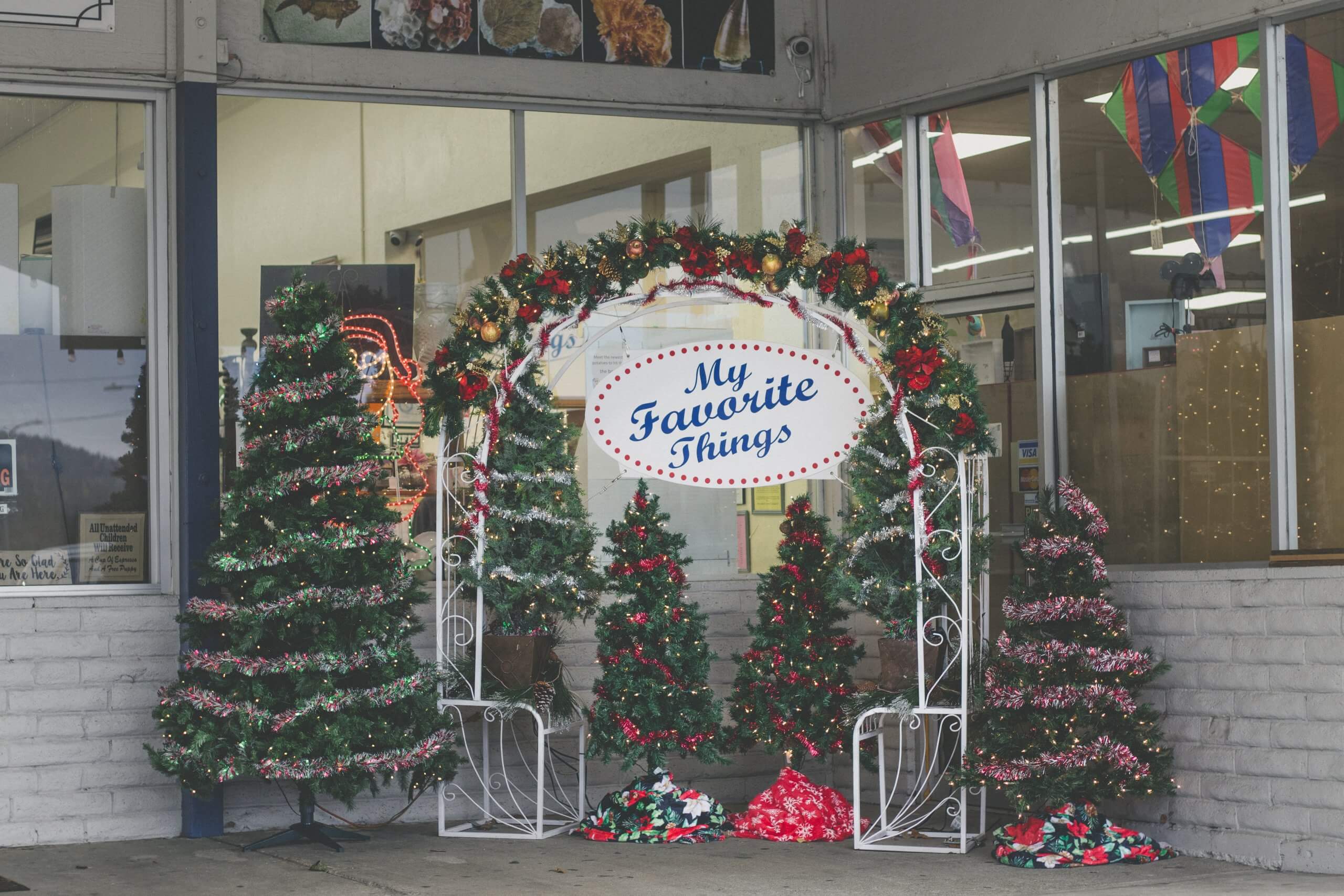 Several Christmas trees adorned with lights and tinsel surrounding an arched garland stand with a sign that says My Favorite Things in front of the My Favorite Things Shop.