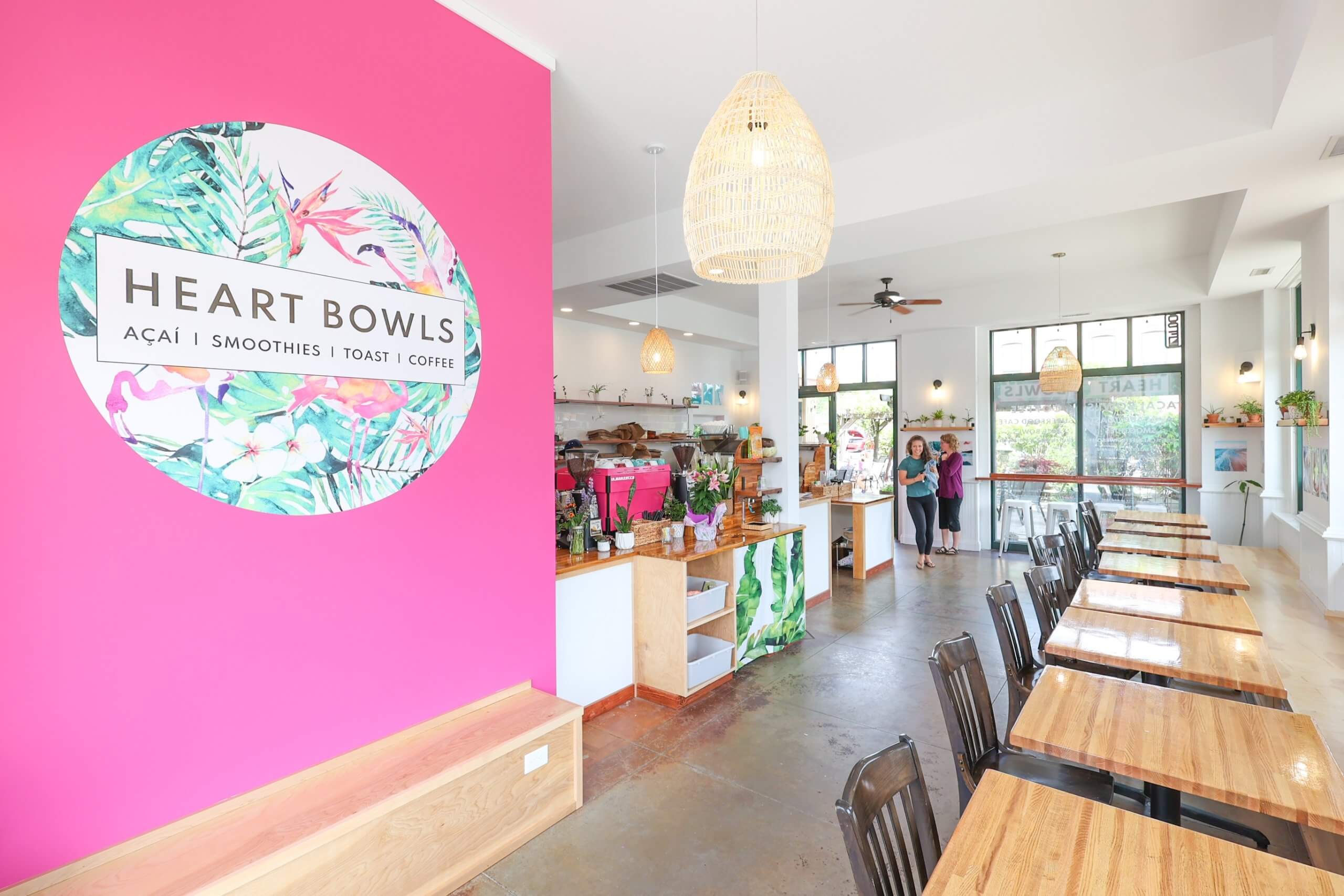 The interior of the Heart Bowls restaurant, showing a row of tables and chairs and a pink wall with the Heart Bowls logo in the foreground and two people waiting at the counter for their orders in the background.