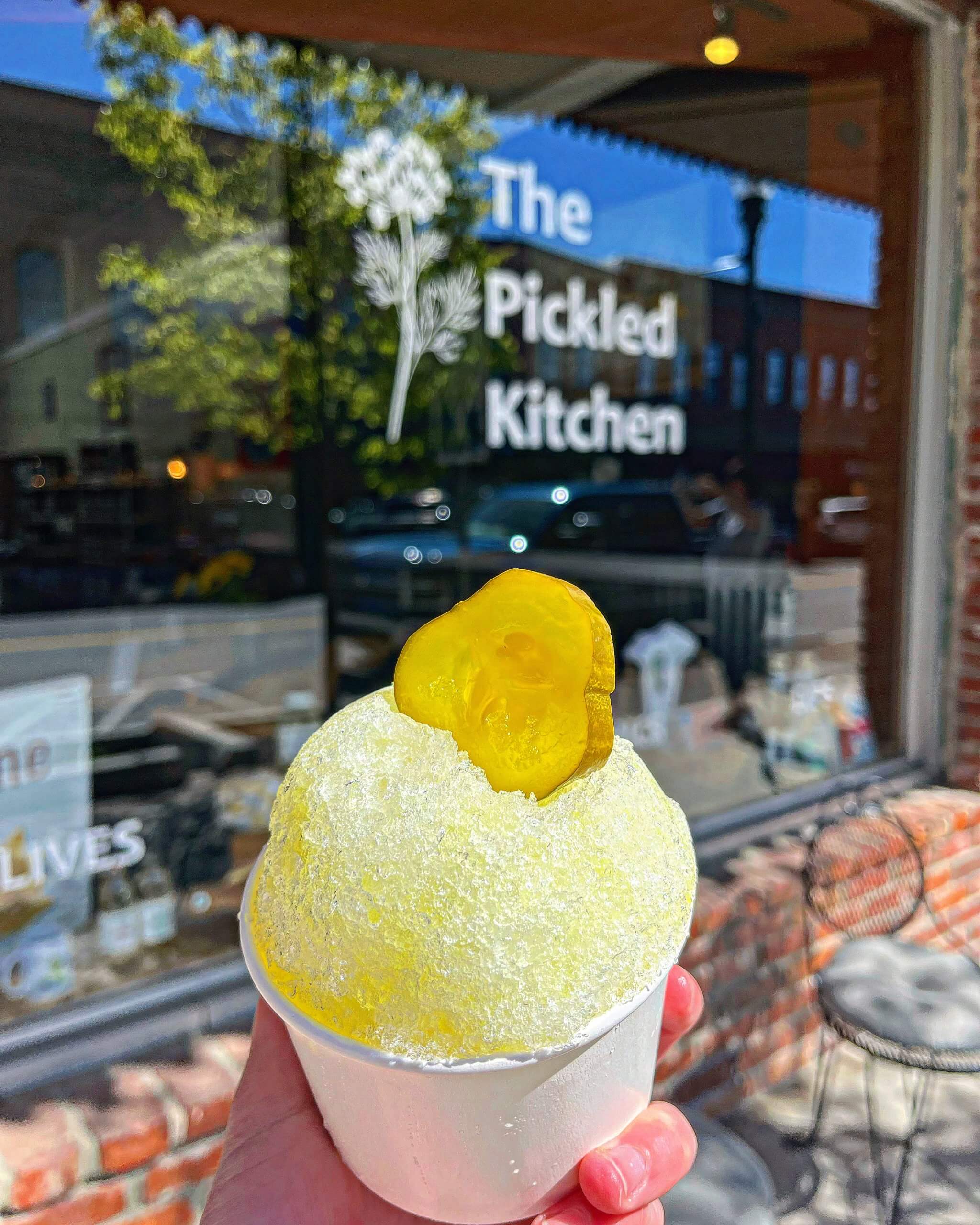 A person's hand holding a pickle-flavored shaved ice topped with a pickle slice in front of the window at The Pickled Kitchen.