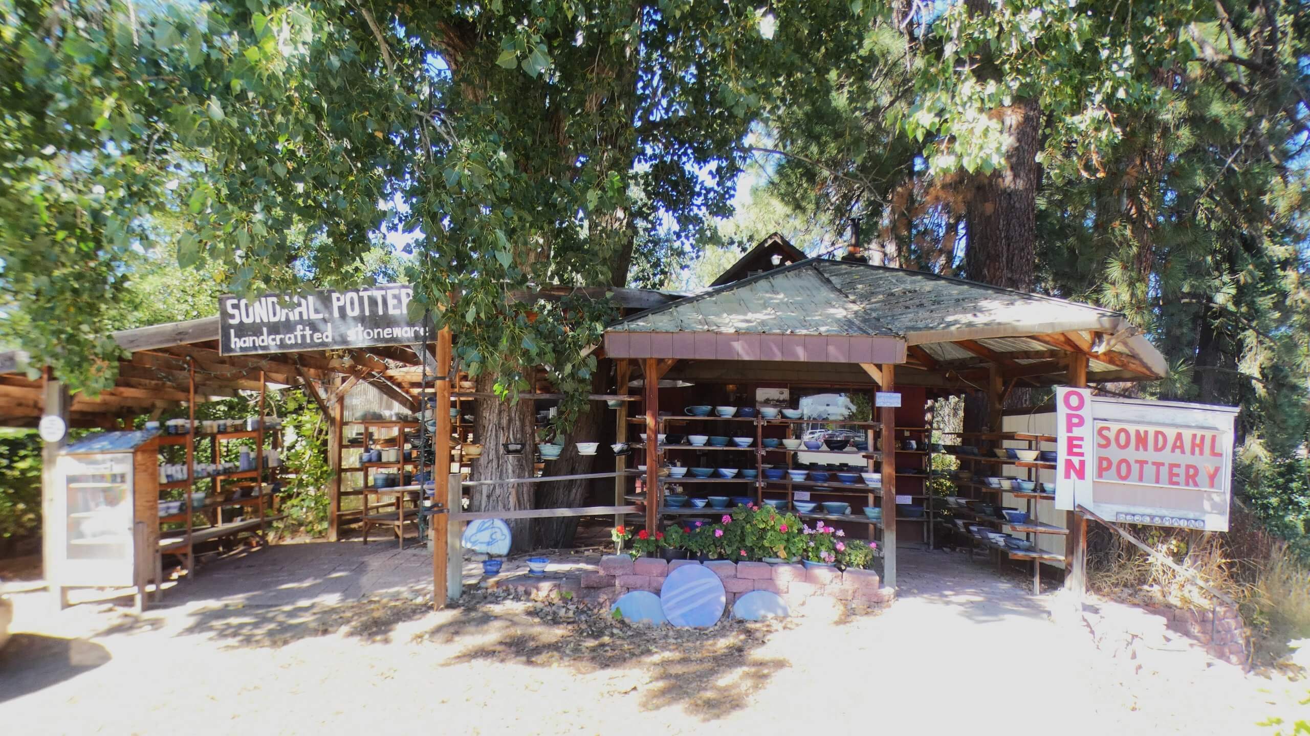 An outside view of the open-air Sondahl Pottery shop surrounded by tall trees with several shelves of potter on display.