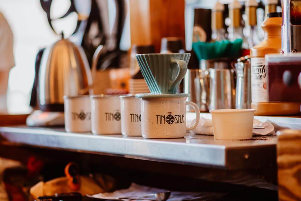 Four tin cups with the Tin Snug logo on them in the foreground, and various coffee pots and bottles of syrups in the background at The Tin Snug.