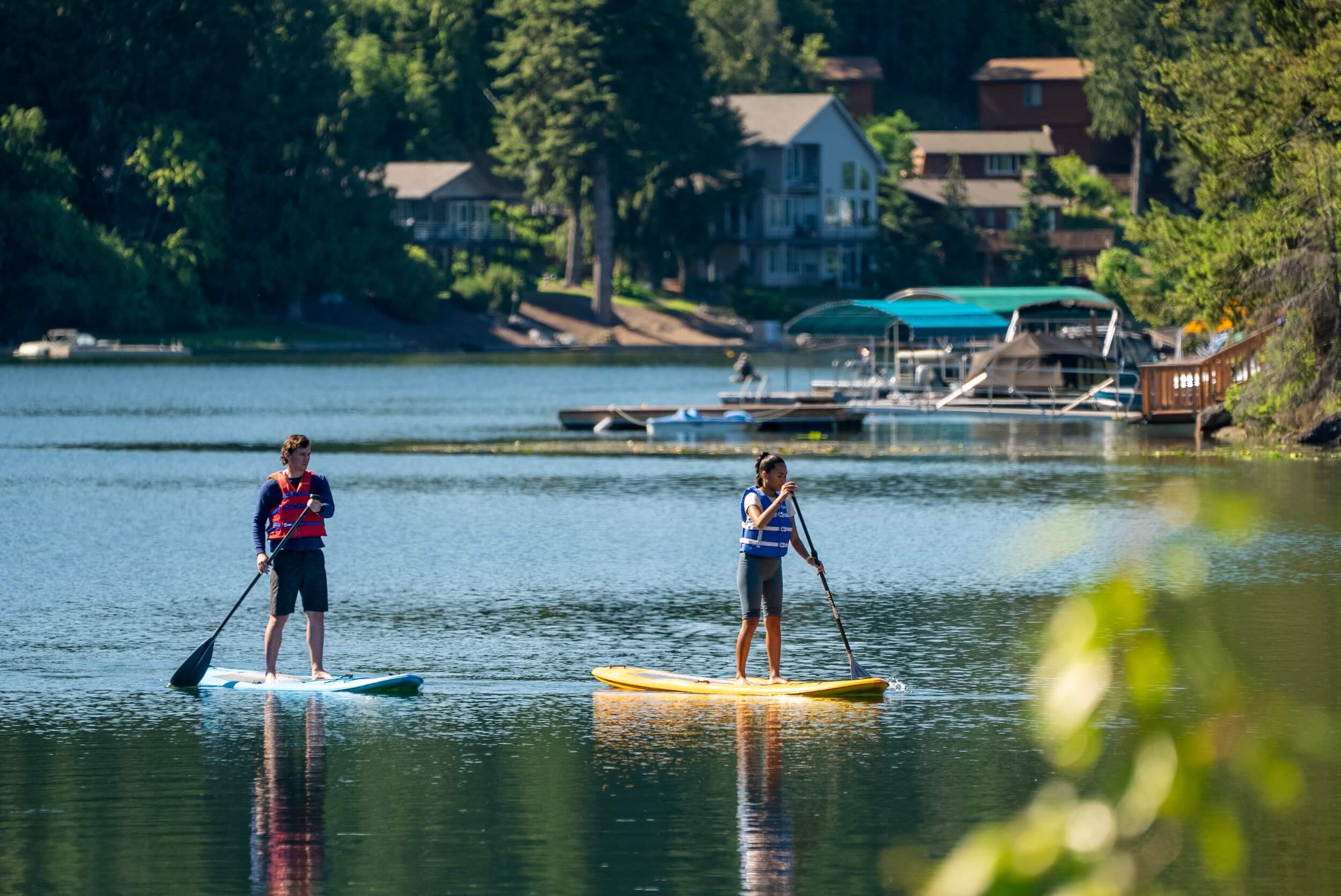 Two people paddleboarding on Spirit Lake in front of a boat dock, and a shore lined with trees and houses in the background.