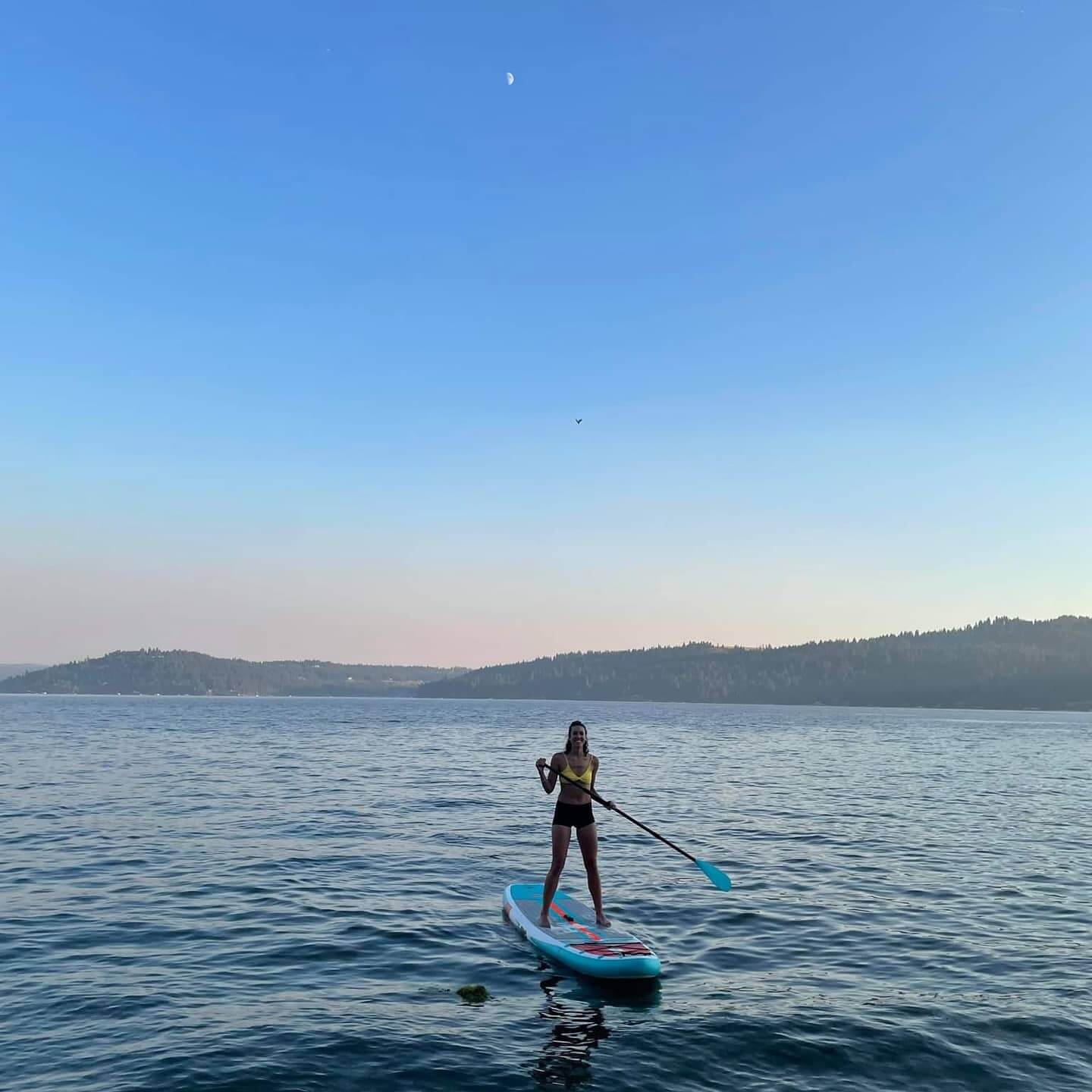 A woman paddleboarding on Lake Coeur d'Alene and tree-covered hills in the background.