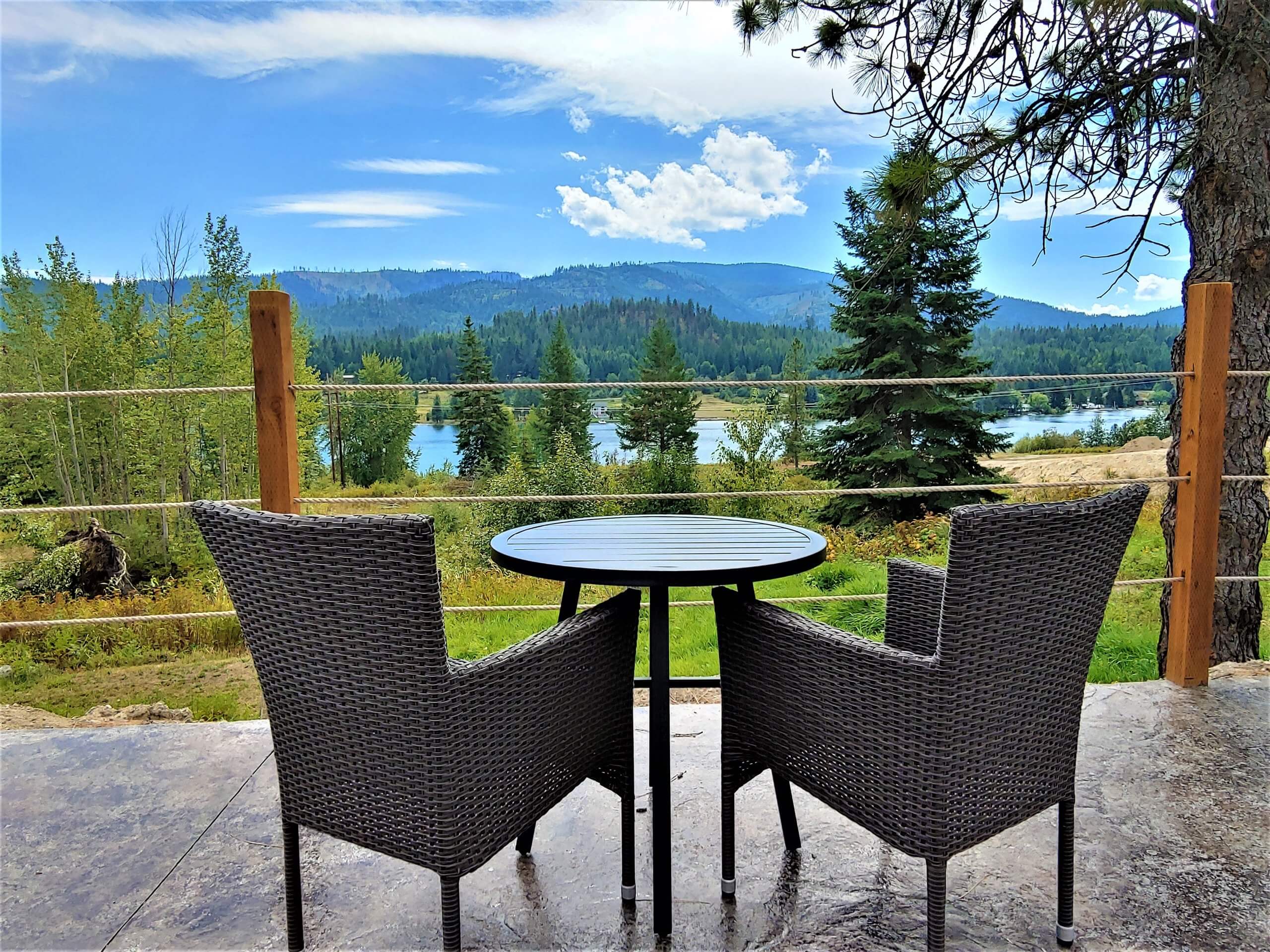 Patio at Eagles Nest Motel with two wicker chairs and a table overlooking a scenic view of trees, a lake, and distant mountains under a blue sky.