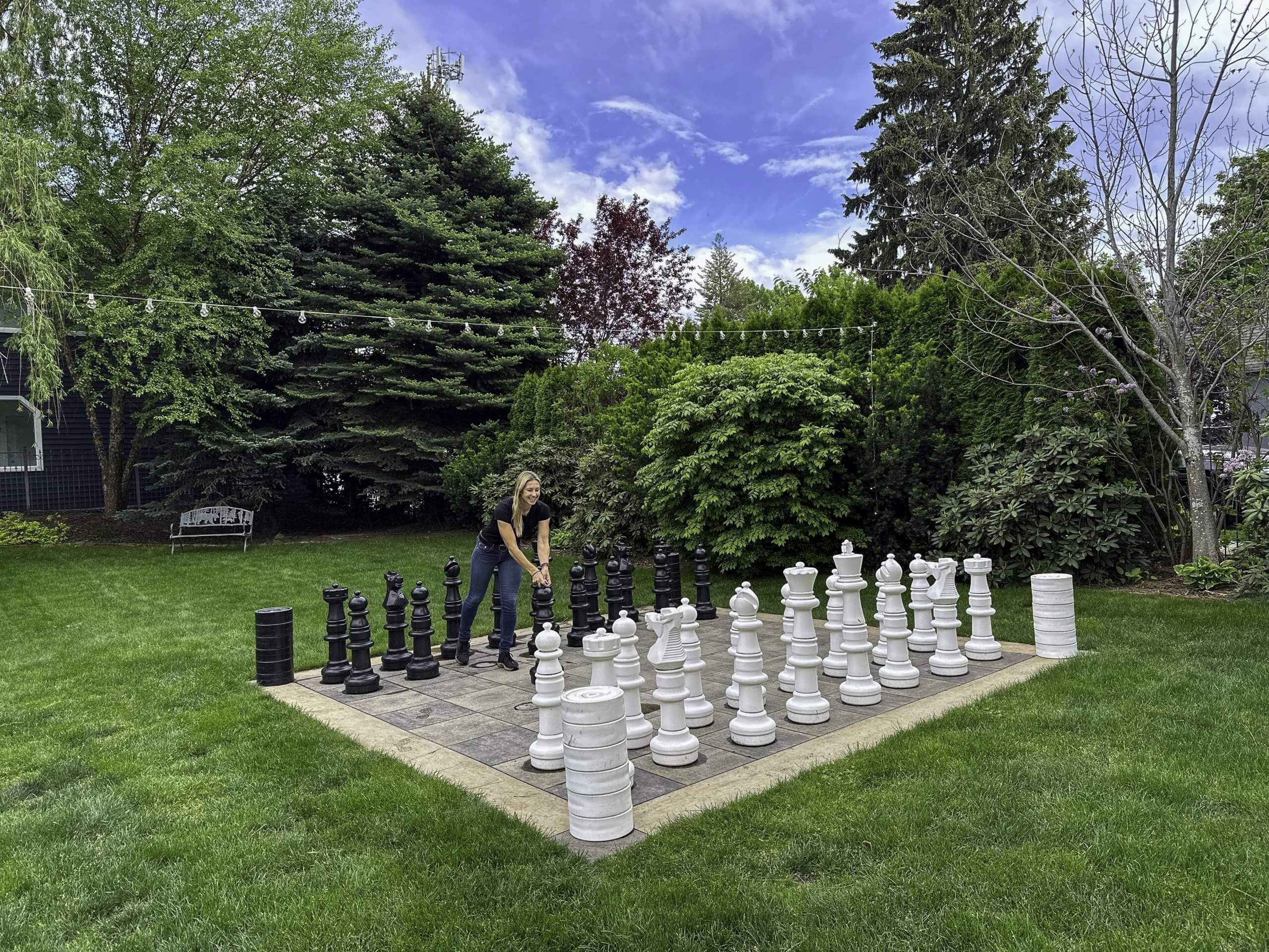 A person playing on a large outdoor chess set at the Roosevelt Inn, surrounded by lush greenery and trees under a blue sky.