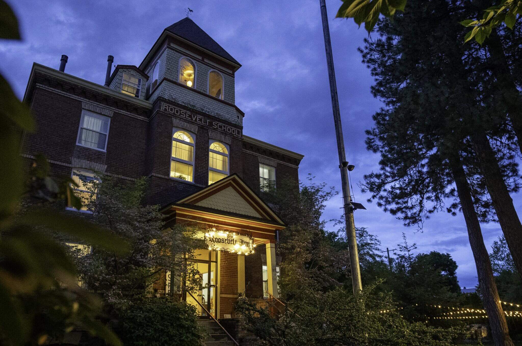 The Roosevelt Inn, a historic building, illuminated at dusk with warm lights, surrounded by tall trees and lush greenery.