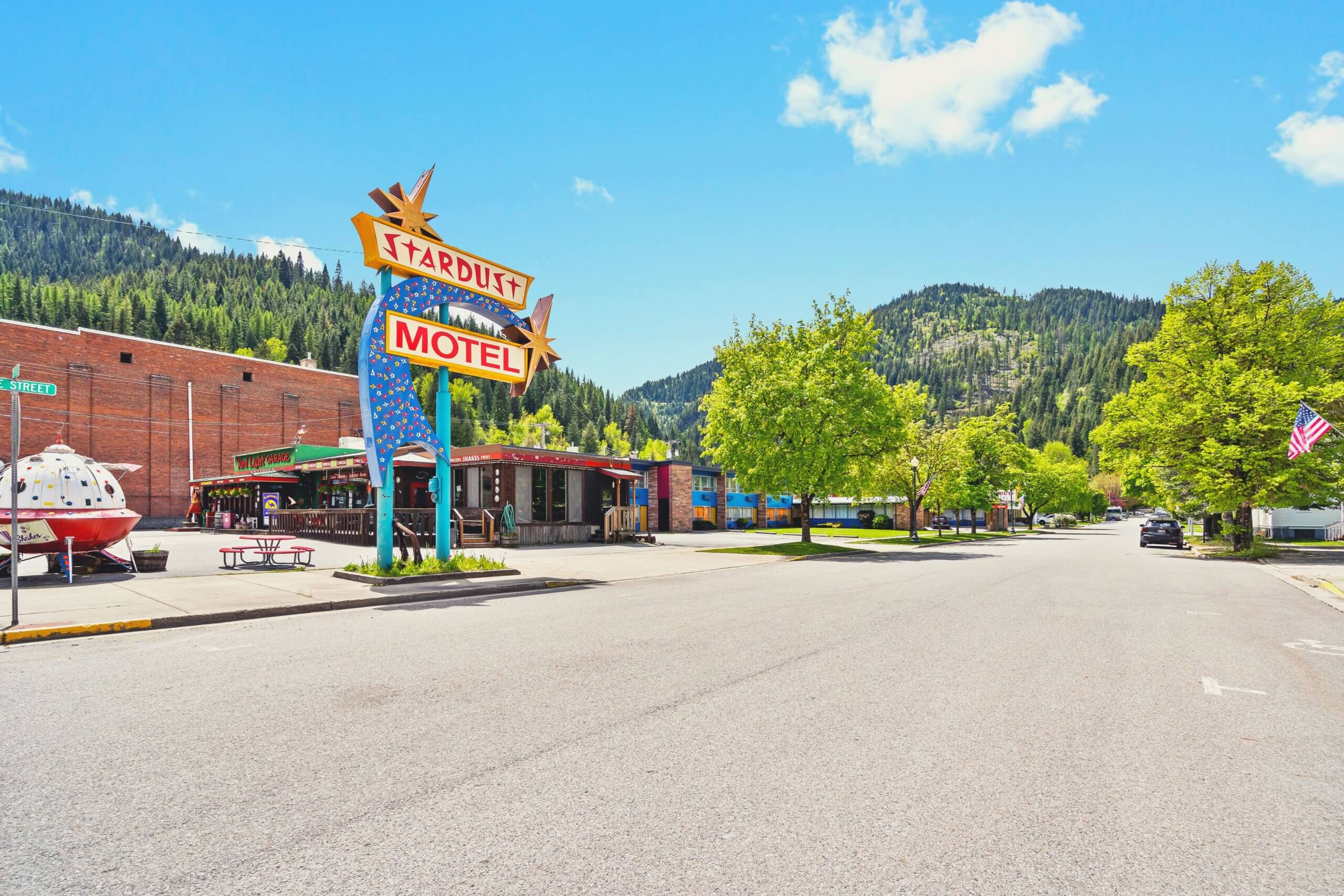 The Stardust Motel in a small mountain town, featuring a retro-style neon sign with a starburst design. The surrounding area includes brick buildings, trees, and a view of forested hills under a bright blue sky.