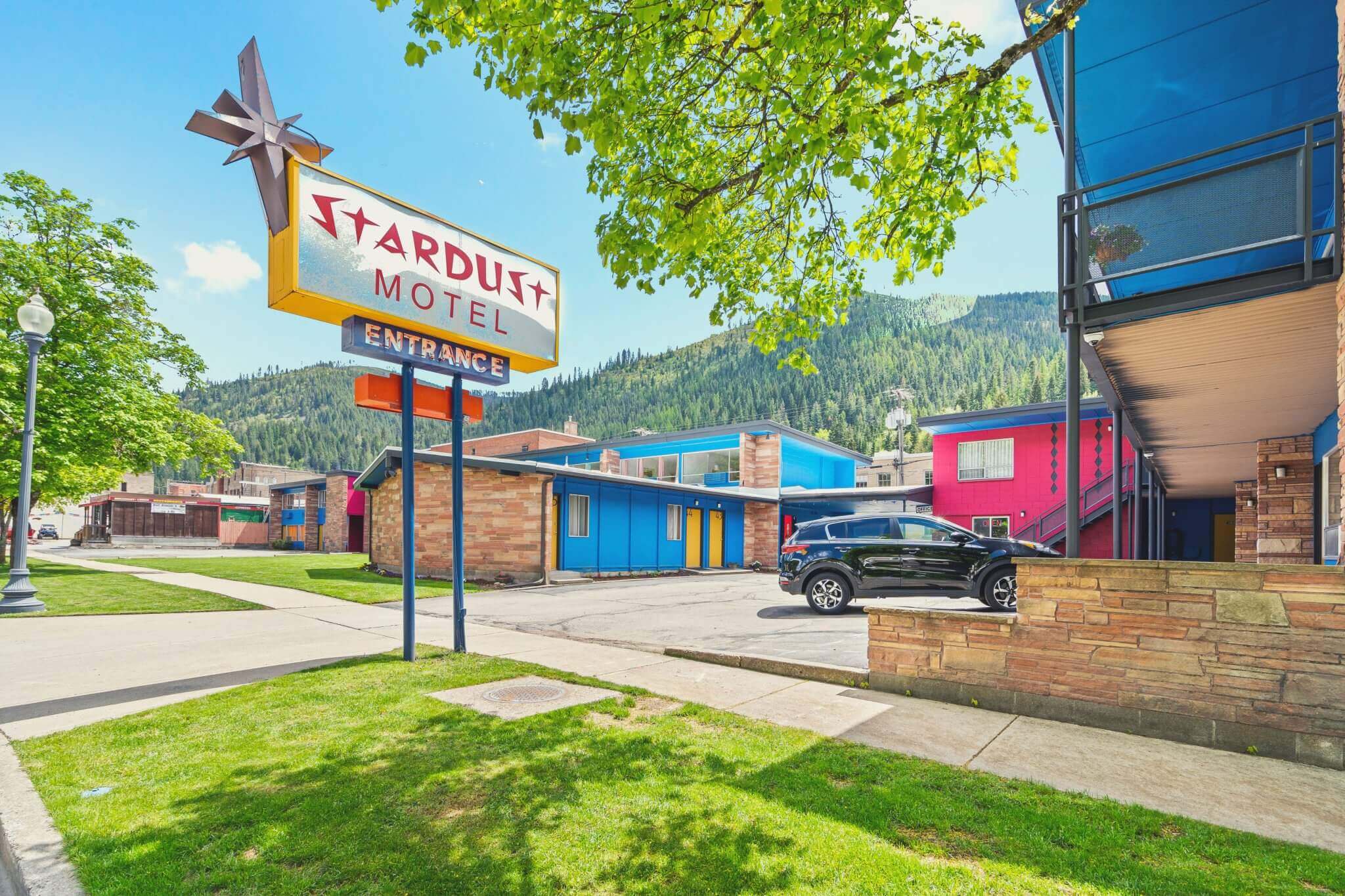 The entrance sign of the Stardust Motel, showcasing a retro design with bold lettering and a starburst motif. The building features vibrant colors and a mid-century modern style, set against a backdrop of green trees and mountain scenery.