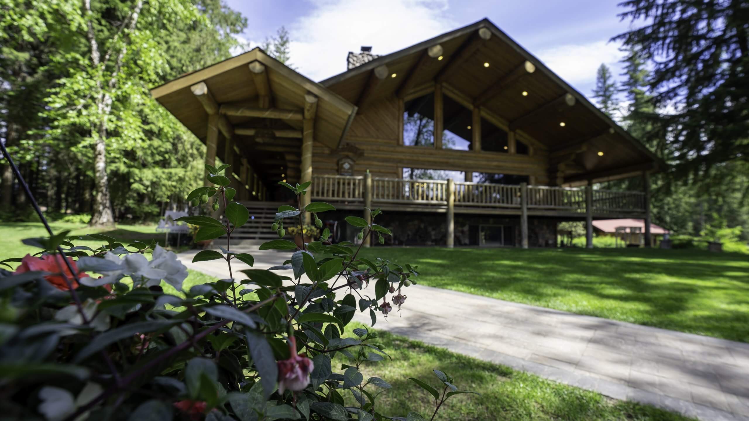 Western Pleasure Ranch, a spacious log cabin with a large porch, surrounded by a lush green forest. Flowering plants are visible in the foreground, adding to the cabin's natural and cozy atmosphere.