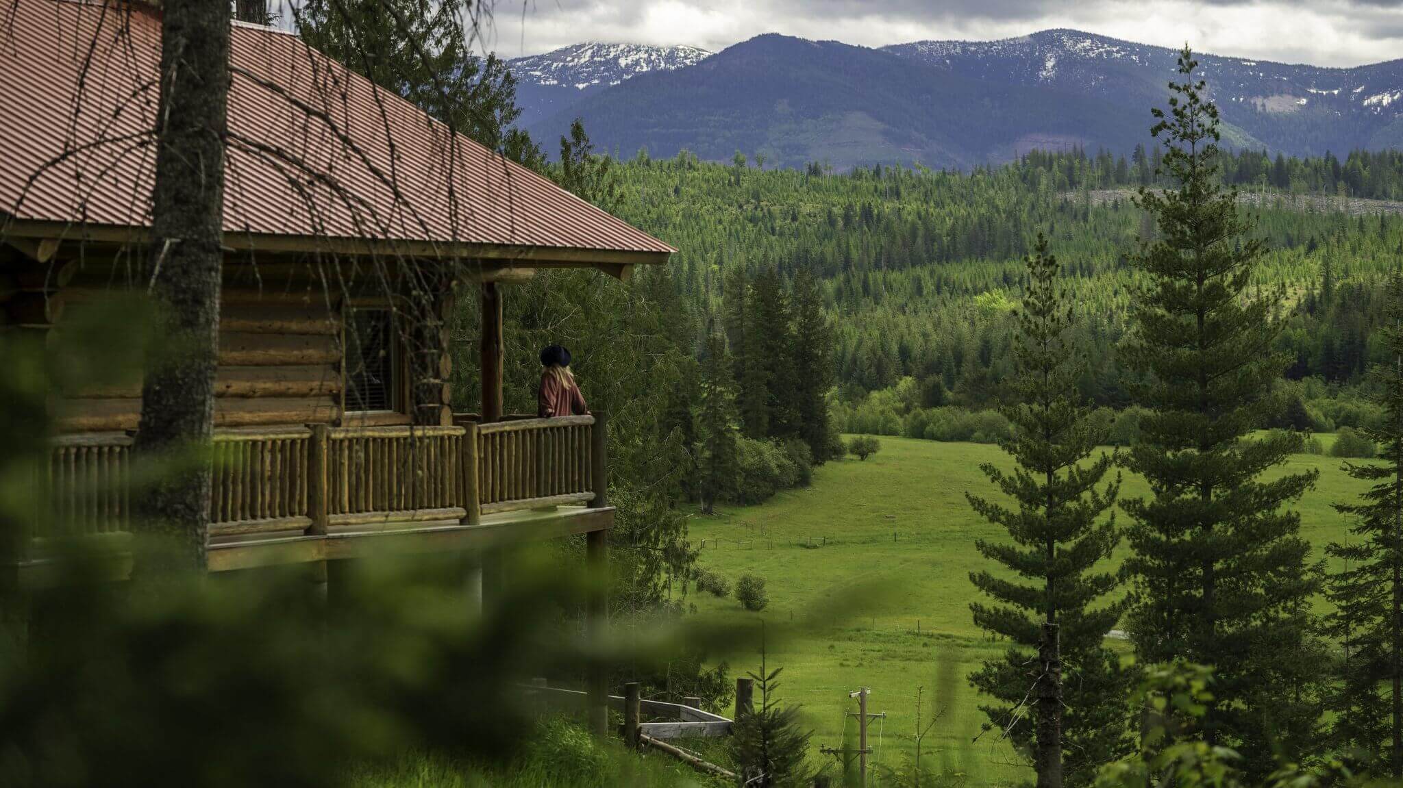 A person standing on the porch of Western Pleasure Ranch with a red roof, overlooking a lush green valley surrounded by pine trees and distant mountains.