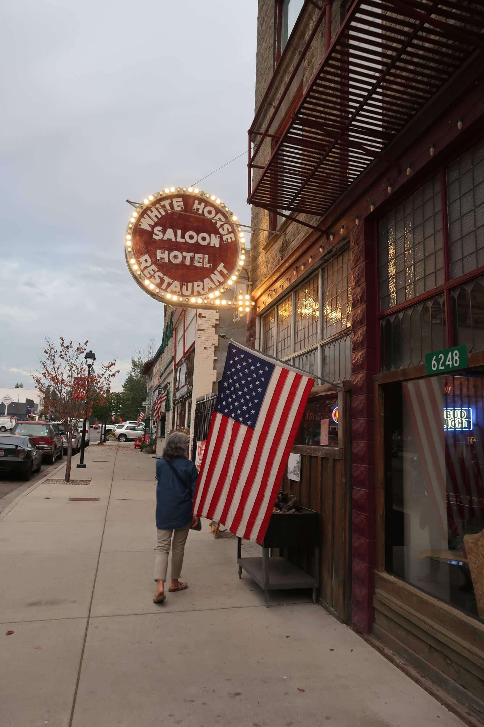 A person standing in front of the historic White Horse Saloon in downtown Spirit Lake, holding an American flag on a cloudy day. The saloon's bright neon sign lights up the street.